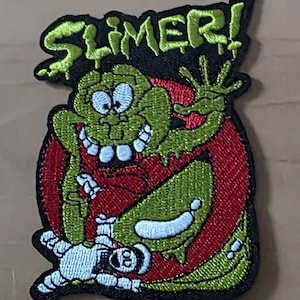 Könnte beinhalten: Gestickter Aufnäher mit der Figur "Slimer" aus Ghostbusters. Der Aufnäher hat ein grünes, rotes und gelbes Farbschema. Das Wort "SLIMER!" ist oben in Grün gestickt. Die Figur wird mit einem breiten Grinsen und ausgestreckter Hand dargestellt.