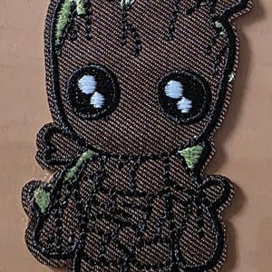 Könnte beinhalten: Gestickter Aufnäher mit einer Cartoonfigur mit brauner, denimartiger Textur. Die Figur hat große, schwarze Augen mit weißen Highlights und grüne Akzente auf Kopf und Armen. Der Aufnäher ist schwarz umrandet.