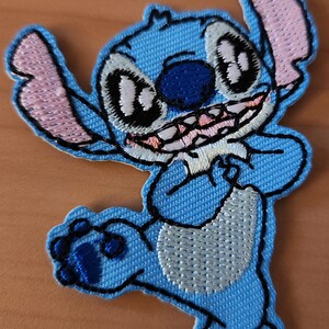 Può includere: Toppa ricamata raffigurante il personaggio di Stitch di Lilo & Stitch. La toppa è principalmente azzurra con contorni neri e dettagli rosa, bianchi e blu scuro. Stitch è raffigurato sorridente con le braccia alzate.