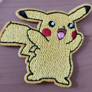 May include: Embroidered yellow Pikachu patch with black-tipped ears, red cheeks, and a smiling expression. The patch has a yellow border and is designed for application on clothing or accessories.