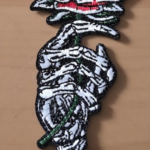 Könnte beinhalten: Gestickter Aufnäher mit einer roten Rose mit schwarzen und weißen Details. Der Stiel und die Blätter sind grün. Das Design beinhaltet einen skelettartigen Stiel und Blätter, die eine einzigartige gotische Ästhetik schaffen.