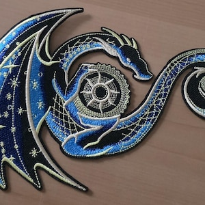Puede incluir: Parche bordado con un dragón estilizado en tonos azules, negros y blancos. El dragón tiene grandes alas con detalles de estrellas y cruces. El diseño incluye un elemento circular en el centro y una cola enrollada.