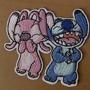 May include: Embroidered patches of Stitch and Angel from Lilo & Stitch. Stitch is blue and smiling, Angel is pink and covering her face. Both have white outlines. The patches are set against a brown background.