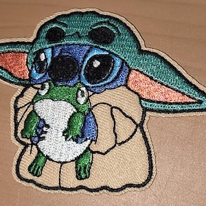 Könnte beinhalten: Gestickter Aufnäher mit einer blau-grünen Figur mit großen Augen, die einen grünen Hut trägt und einen grün-weißen Frosch hält. Der Aufnäher hat einen beigen Rand, geeignet für Kleidung oder Accessoires.