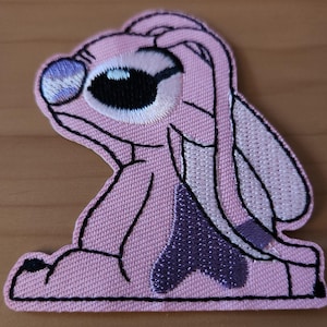 Könnte beinhalten: Ein rosa gestickter Aufnäher mit einer Cartoonfigur mit großen Augen und langen Ohren. Der Aufnäher hat eine schwarze Umrandung und einen lila Akzent. Das Auge der Figur ist schwarz und weiß, mit einem lila Akzent.