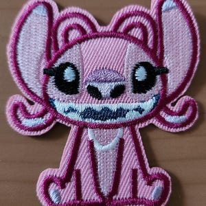 Könnte beinhalten: Ein pinkfarbener, gestickter Aufnäher mit einer Cartoonfigur mit großen Ohren, schwarzen Augen und einem breiten Grinsen. Der Aufnäher hat eine weiße Umrandung und ist zum Aufbügeln oder Aufnähen auf Stoff gedacht. Die Figur sitzt.