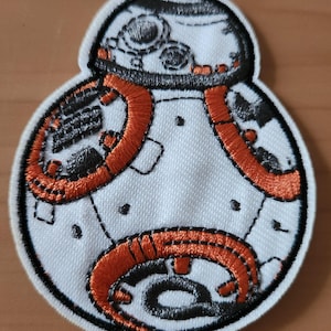 Könnte beinhalten: Gestickter Aufnäher mit dem BB-8-Droiden aus Star Wars. Der Aufnäher hat einen weißen Hintergrund mit orangefarbenen und schwarzen Stickdetails. Das Design umfasst den Kopf und den Körper des Droiden mit aufwendigen Nähten.