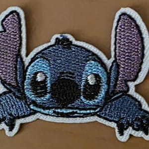 Könnte beinhalten: Gestickter Aufnäher mit der Figur Stitch aus Lilo & Stitch. Der Aufnäher hat einen blauen Körper, große lila Ohren und weiße Akzente. Das Design ist detailliert mit schwarzen Umrissen und weißen Augen.