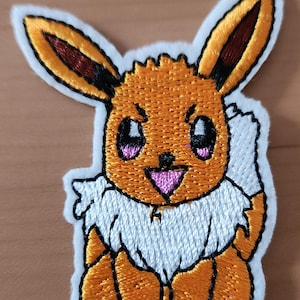 Könnte beinhalten: Gestickter Aufnäher mit dem Eevee-Charakter, mit orangefarbenem Fell, großen Ohren und einem weißen, flauschigen Kragen. Der Aufnäher hat einen weißen Rand und ist für das Aufnähen auf Kleidung oder Accessoires bestimmt.