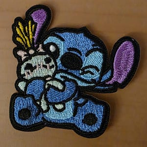 May include: Embroidered patch featuring the character Stitch from Lilo & Stitch, in shades of blue, purple, and black. Stitch is hugging a small, light blue and white creature. The patch has a black border and is on a light brown surface.