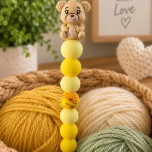 Puede incluir: Un ganchillo con un adorno de oso de color marrón claro y un mango decorado con una pila vertical de siete cuentas esféricas en varios tonos de amarillo, incluida una cuenta jaspeada amarilla y naranja, colocado verticalmente en una cesta.