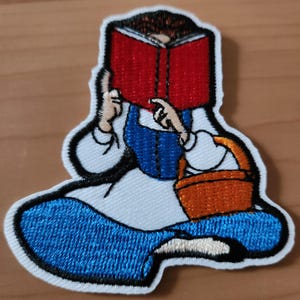 Può includere: Toppa ricamata raffigurante un personaggio dei cartoni animati che legge un libro. Il personaggio indossa un vestito blu e tiene un cestino arancione. Il libro è rosso e la toppa ha un bordo bianco con cuciture nere.