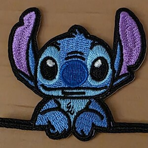 Könnte beinhalten: Gestickter Aufnäher mit der Figur Stitch aus Lilo & Stitch. Der Aufnäher hat einen blauen Körper, lila Ohren und schwarze Umrisse. Die Figur lächelt mit großen schwarzen Augen und einer blauen Nase. Der Aufnäher befindet sich auf einer hellbraunen Oberfläche.