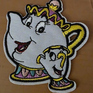 Könnte beinhalten: Gestickter Aufnäher mit einer Cartoon-Teekanne und -Tasse mit lächelnden Gesichtern. Die Teekanne ist hellgrau mit gelben, rosa und schwarzen Akzenten. Die Tasse ist hellgrau mit gelben und schwarzen Akzenten. Der Aufnäher ist für den Stoffaufnäher bestimmt.