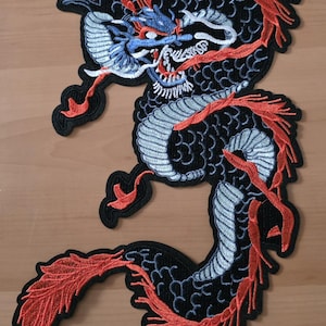 Könnte beinhalten: Gestickter Aufnäher mit einem Drachendesign. Der Drache hat einen schwarzen Körper mit hellblauen Schuppen, roten Akzenten und einem blau-weißen Kopf. Geeignet für Kleidung oder Accessoires.