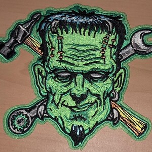Könnte beinhalten: Gestickter Aufnäher mit einem grünen Frankenstein-Monster mit schwarzen Haaren, Stichen und einem Ziegenbart. Ein Hammer und ein Schraubenschlüssel sind hinter dem Kopf des Monsters gekreuzt. Der Aufnäher hat einen grünen Rand.