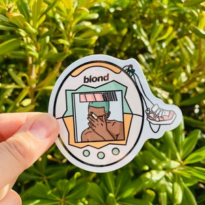 May include: A white, yellow, and green sticker of a digital pet toy with a picture of Frank Ocean on it. The toy has the word "blond" on it and a picture of a shoe on the side.