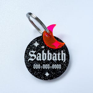 May include: A black glittery pet tag with the name "Sabbath" in white gothic font and a phone number. A neon pink crescent moon charm and a silver ring are attached to the tag. The tag has two white star designs.
