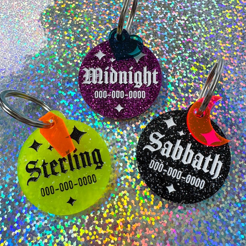 May include: Three glittery pet ID tags with silver key rings. One is purple with "Midnight" in white script, another is black with "Sabbath", and the third is neon green with "Sterling". Each tag has a phone number.