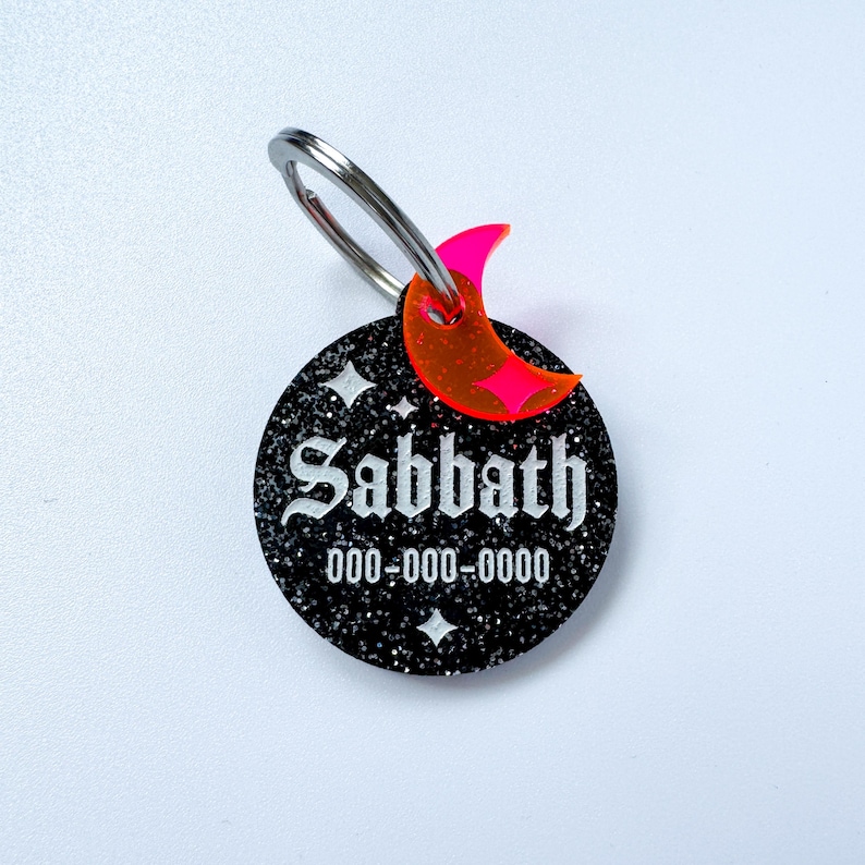 May include: A black glittery pet tag with the name "Sabbath" in white gothic font and a phone number. A neon pink crescent moon charm and a silver ring are attached to the tag. The tag has two white star designs.