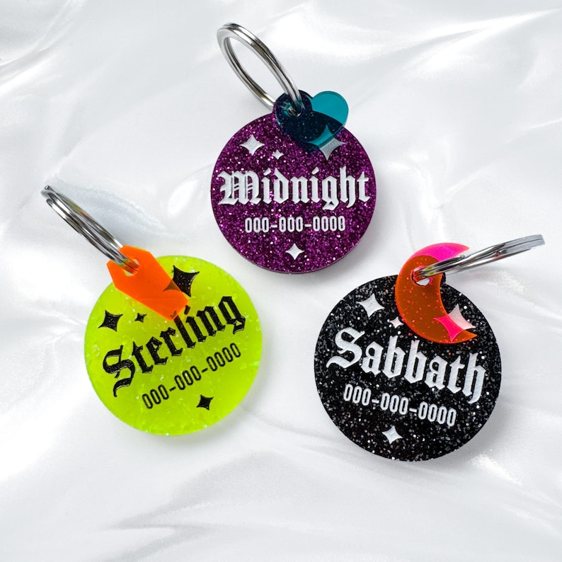 May include: Three round pet tags with silver key rings. One tag is purple with the name "Midnight", another is neon green with "Sterling", and the third is black with "Sabbath". Each tag has a phone number.