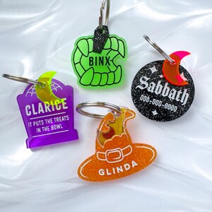 May include: Four Halloween-themed keychains. One is a green set of fangs with "BINX" text. Another is a purple tombstone with "CLARICE" and "IT PUTS THE TREATS IN THE BOWL" text. A third is a black circle with "Sabbath" and a phone number. The last is an orange witch hat with "GLINDA" text.
