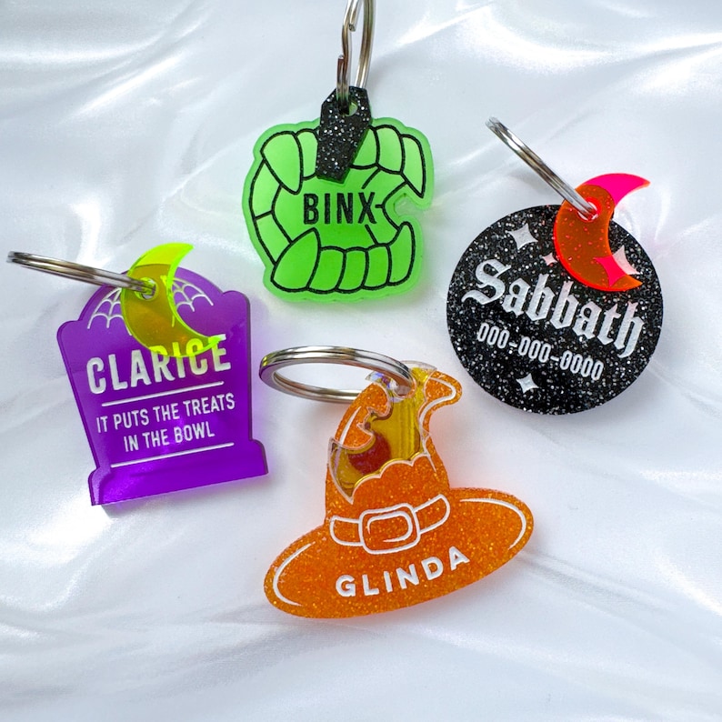 May include: Four Halloween-themed keychains. One is a green set of fangs with "BINX" text. Another is a purple tombstone with "CLARICE" and "IT PUTS THE TREATS IN THE BOWL" text. A third is a black circle with "Sabbath" and a phone number. The last is an orange witch hat with "GLINDA" text.