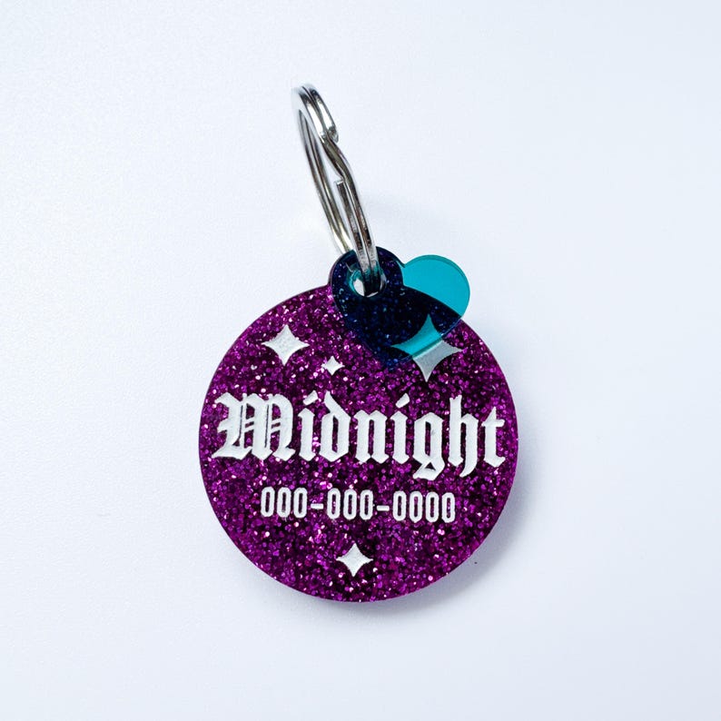 May include: A round, glittery purple pet tag with the name "Midnight" in white, stylized lettering. The tag includes a phone number and starburst designs. It is attached to a silver keyring with a teal heart-shaped charm.
