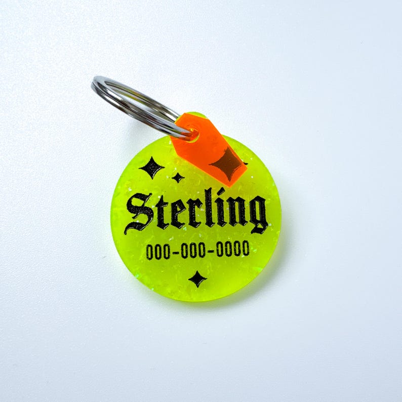 May include: A neon green pet tag with the name "Sterling" in black gothic font and a phone number. The tag has a silver ring and an orange accent piece. Four black starbursts are also on the tag.