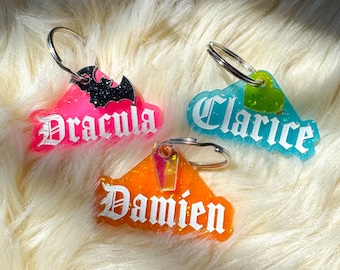 Pode incluir: Três chaveiros coloridos com argolas prateadas. Um é rosa com o nome "Dracula" e um morcego, outro é azul com "Clarice", e o terceiro é laranja com "Damien". Todos têm detalhes brilhantes.