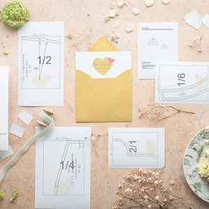 May include: A flat lay of sewing patterns for pants and a jacket. The patterns are printed on white paper and include instructions for cutting and sewing. The patterns are arranged on a light pink surface with a yellow envelope, a spool of thread, and white flowers.