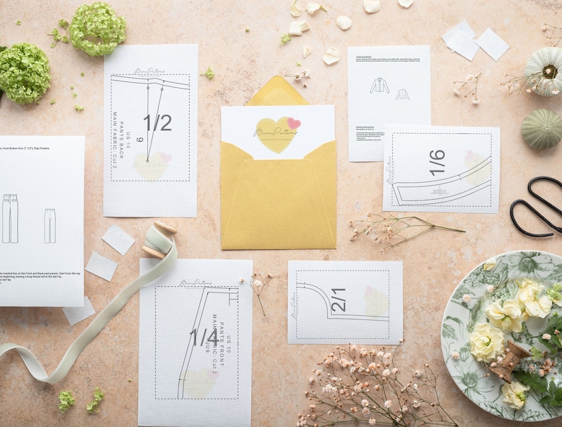 May include: A flat lay of sewing patterns for pants and a jacket. The patterns are printed on white paper and include instructions for cutting and sewing. The patterns are arranged on a light pink surface with a yellow envelope, a spool of thread, and white flowers.