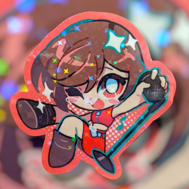 May include: A holographic sticker featuring a cartoon character with brown hair, wearing a red outfit, and holding a microphone. The sticker has a pink border and star accents.