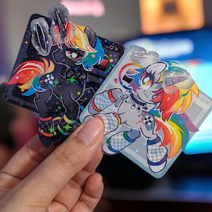 K&ouml;nnte beinhalten: Zwei holografische Aufkleber mit Cartoon-Einhorn-Charakteren mit Regenbogenm&auml;hne und -schwanz. Das Einhorn links ist schwarz mit einem Regenbogen-Karomuster auf den Beinen. Das Einhorn rechts ist grau mit einem Regenbogen-Karomuster auf den Beinen und einem Regenbogenmuster auf dem Schwanz. Beide Aufkleber haben eine glitzernde Oberfl&auml;che.