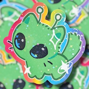 May include: A green alien sticker with a rainbow border. The alien has large black eyes, antennae, and is decorated with star-shaped and diamond-shaped sparkles. The sticker has a holographic effect, creating a shimmering appearance.