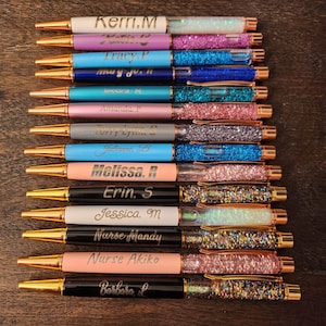 Peut inclure: Un ensemble de 12 stylos personnalisés avec des accents dorés et des barils remplis de paillettes. Les stylos sont disposés verticalement avec les noms de chaque personne gravés sur le baril. Les noms incluent Kerri.M, Tracy.P, Jessica.M, Amanda.P, Julienne.B, Melissa.R, Erin.S, Jessica.M, Nurse Mandy, Nurse Akiko et Barbara.L.
