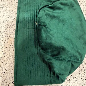 May include: A folded, emerald green fabric item with a quilted, ribbed texture on one side and a smooth, plush texture on the other. The item appears to be a hat or a similar head covering, with a rounded shape and a folded brim.