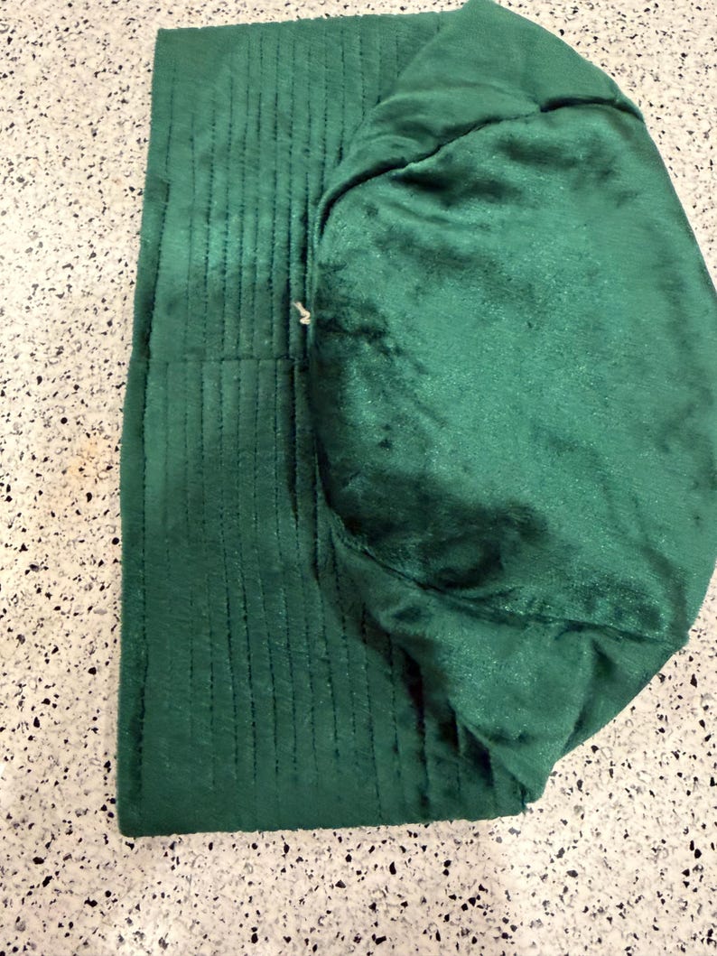 May include: A folded, emerald green fabric item with a quilted, ribbed texture on one side and a smooth, plush texture on the other. The item appears to be a hat or a similar head covering, with a rounded shape and a folded brim.