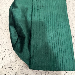 May include: A folded, emerald green fabric item with a textured, ribbed design on one side and a smooth, satin-like finish on the other. The item appears to be a head covering or accessory, possibly a turban or a similar garment.