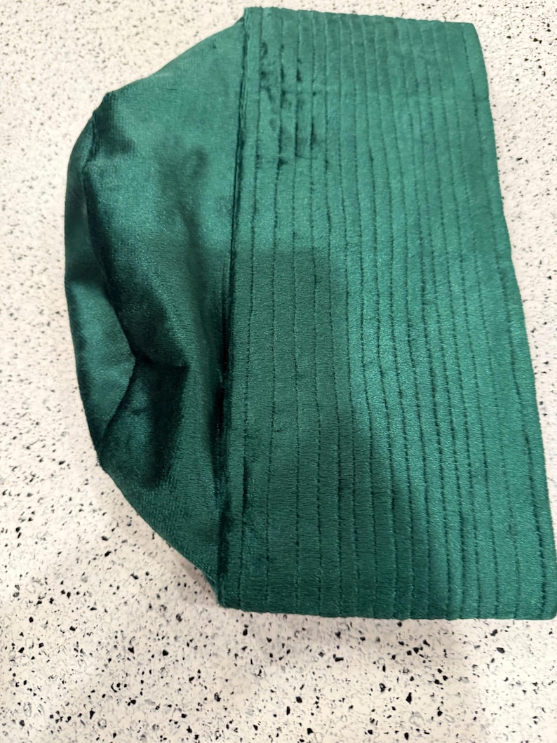 May include: A folded, emerald green fabric item with a textured, ribbed design on one side and a smooth, satin-like finish on the other. The item appears to be a head covering or accessory, possibly a turban or a similar garment.