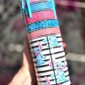 May include: A tumbler with a blue, pink, and glitter design. The top has a blue drip effect. The middle section features alternating pink and blue glitter stripes. The bottom section has a black and white striped pattern with blue cartoon character illustrations.