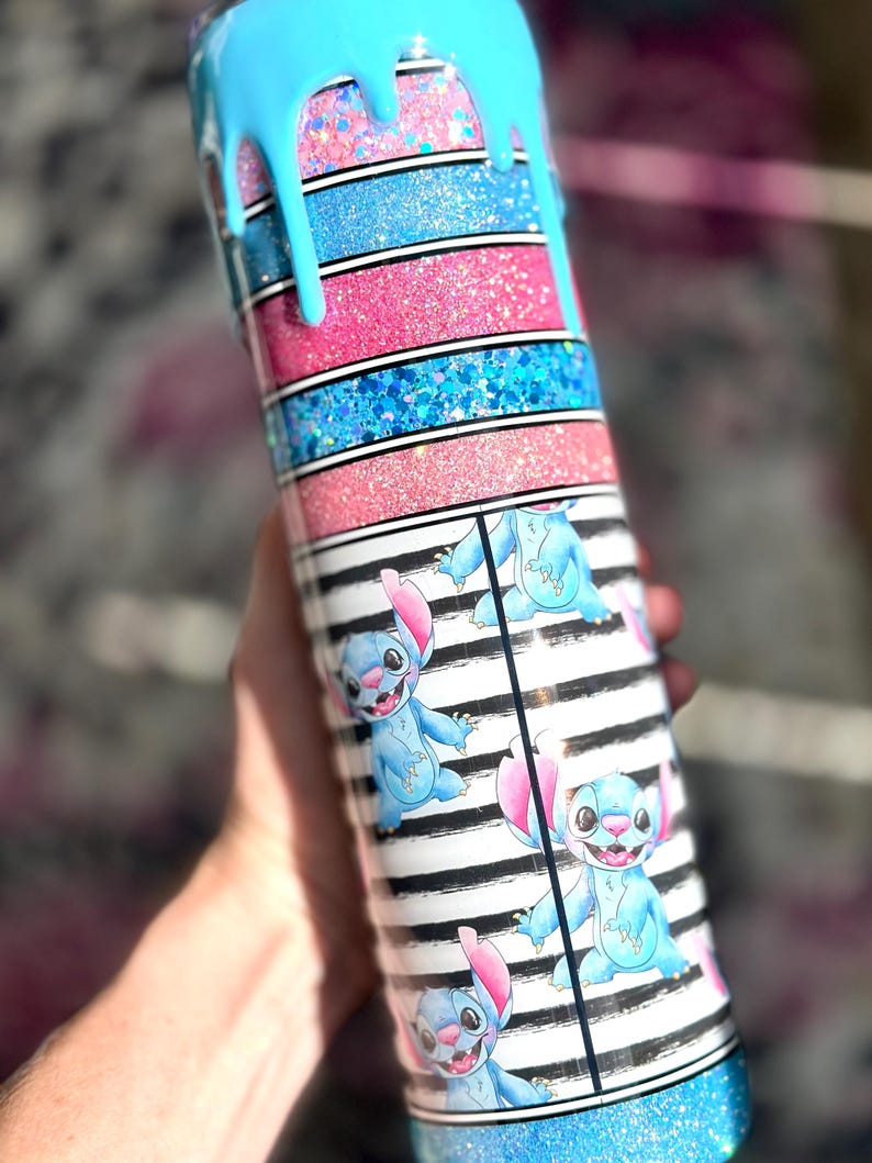 May include: A tumbler with a blue, pink, and glitter design. The top has a blue drip effect. The middle section features alternating pink and blue glitter stripes. The bottom section has a black and white striped pattern with blue cartoon character illustrations.