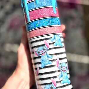 May include: A glittery tumbler with a blue, pink, and black and white striped design. The top has a blue drip effect. The tumbler features cartoon images of Stitch.