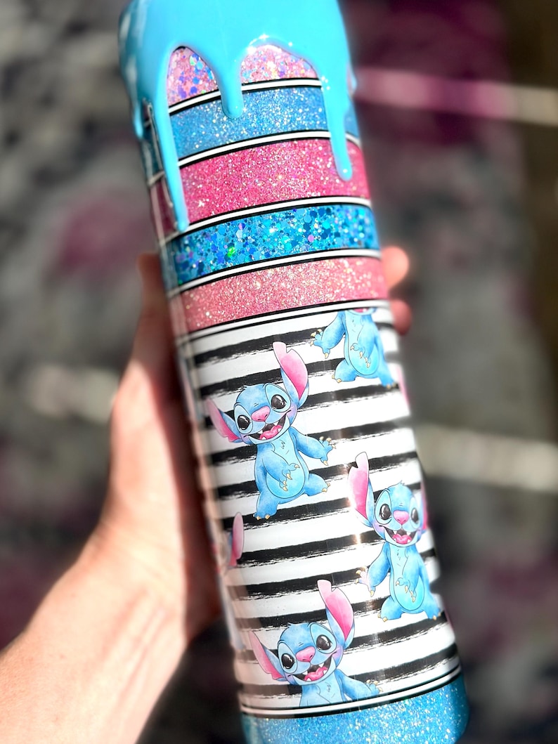 May include: A glittery tumbler with a blue, pink, and black and white striped design. The top has a blue drip effect. The tumbler features cartoon images of Stitch.