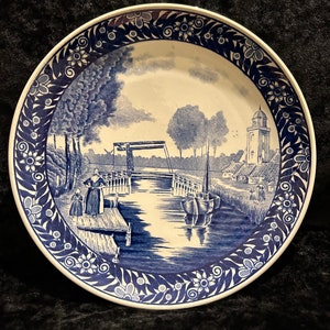 May include: A blue and white ceramic plate featuring a Dutch scene of a canal, bridge, and houses. The plate has a decorative border of blue flowers and leaves.