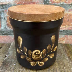 May include: A black ceramic jar with a wooden lid and a gold floral design. The jar features a stylised rose and leaves.