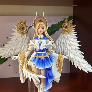 May include: A white and gold figurine of a woman with long blonde hair wearing a white and blue dress and a golden crown. She has large white wings with gold accents. The figurine is standing on a black base.