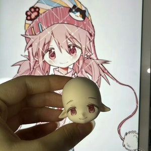 May include: A light pink and white doll head with pink eyes and a closed mouth smile. The doll head has pointed ears and is being held by a hand.