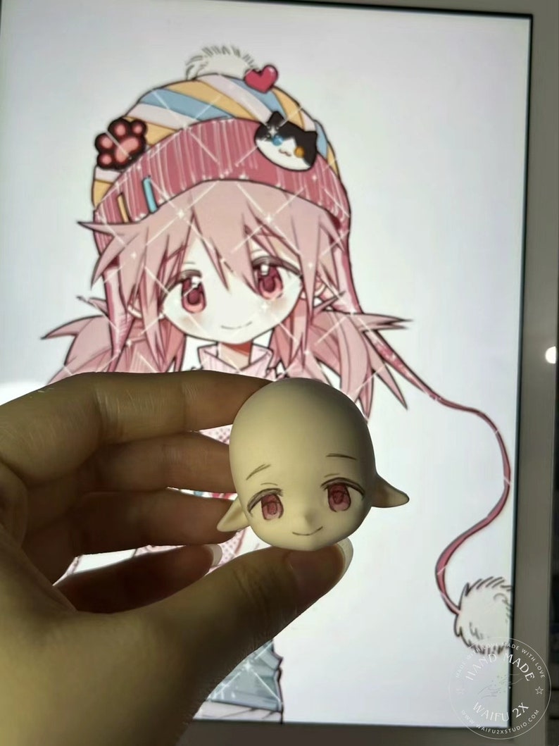 May include: A light pink and white doll head with pink eyes and a closed mouth smile. The doll head has pointed ears and is being held by a hand.