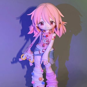 May include: A pink and white anime figurine with pink hair, wearing a pink and white shirt with a cat design, blue denim shorts, and pink and white socks with colorful pom-poms. The figurine is holding a pink and white plush toy. The figurine is standing on a black base with a white sticker that says "Hand Made with Love Made Without Love" and "Waifu 2X".